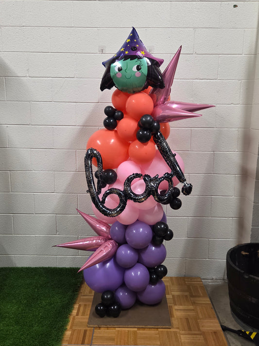 Witch-themed Halloween balloon arrangement featuring a large balloon witch hat, black and purple balloons, green accents, and spooky Halloween decorations like bats and cauldrons.